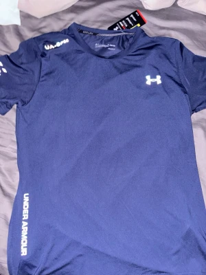 Blå träningströja från Under Armour - Snygg mörkblå träningströja från Under Armour med vit logga på bröstet och axeln samt text längs sidan. Tillverkad i ett lätt och snabbtorkande funktionsmaterial, perfekt för gym eller löpning. Kortärmad och med rund hals.