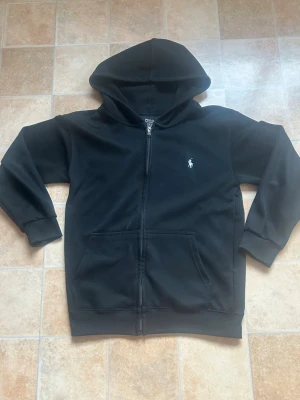Svart zip hoodie från Polo Ralph Lauren - Svart hoodie med dragkedja från Polo Ralph Lauren. Klassisk design med huva, två stora fickor framtill och den ikoniska vita logon broderad på bröstet. Tillverkad i mjukt material som känns skönt mot huden. Perfekt för en chill och stilren look.