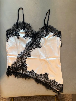 Vit spets med svart spets - Snygg topp i vit satin med tunna axelband och dekorativa svarta spetsdetaljer längs kanterna. Asymmetrisk design med spets både upptill och nertill ger en lyxig och trendig look. Perfekt för dig som gillar eleganta underkläder med det lilla extra.