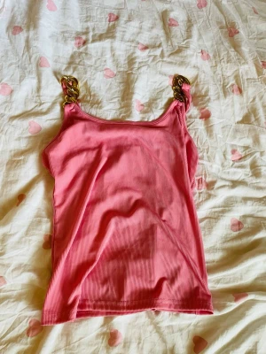 Rosa ribbad linnetopp med guldkedjor - Säljer en rosa ribbad linnetopp med breda axelband i guldfärgad kedja. Toppen har en croppad passform och är perfekt för dig som vill sticka ut lite extra. Materialet känns mjukt och stretchigt, vilket gör den bekväm att bära.