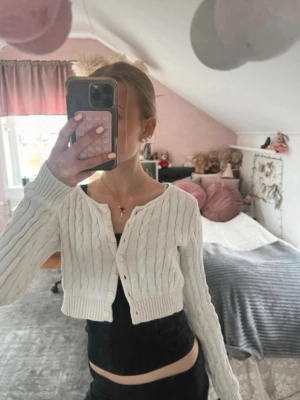 Vit kabelstickad kofta Brandy Melville - Säljer en vit kabelstickad kofta från Brandy Melville. Koftan är croppad med långa ärmar och har knappar framtill. Klassisk kabelstickad design och ribbade muddar vid ärmslut och nederkant. Perfekt att styla över ett linne eller t-shirt.