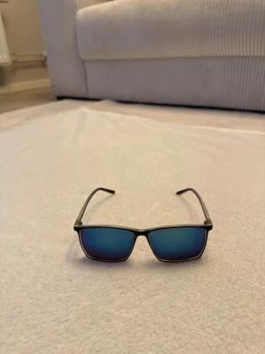 Blåa solglasögon med spegelglas från Jack and Jones  - Snygga solglasögon med svart, rektangulär båge och blå spegelglas. Glasögonen har en modern och stilren look som passar perfekt till sommaren. Lätta bågar i syntetmaterial gör dem bekväma att bära hela dagen.