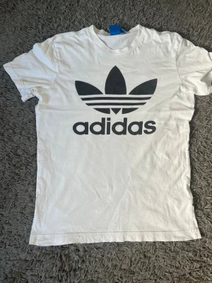 Vit Adidas t-shirt med stor logga - Klassisk vit t-shirt från Adidas med stor svart Trefoil-logga och text på bröstet. T-shirten har rund halsringning och korta ärmar. Tillverkad i mjuk bomull för en bekväm känsla och enkel stil.