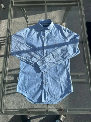Polo Ralph Lauren Skjorta – Ljusblå – Slim Fit – S - Säljer en stilren ljusblå skjorta från Polo Ralph Lauren i modellen "Slim Fit". En tidlös klassiker i 100% bomull som är ett måste i basgarderoben. Den är i storlek S och har den ikoniska broderade loggan på bröstet. Skjortan sitter snyggt och smalt, och passar perfekt att bära både uppknäppt över en t-shirt eller stängd till ett par finbyxor eller jeans. Väldigt fint skick  Kort info:  Märke: Polo Ralph Lauren  Passform: Slim Fit  Storlek: S  Material: 100% bomull  Färg: Ljusblå 