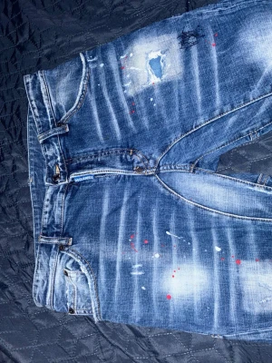 Blå slitna jeans från Dsquared2 - Säljer ett par blå jeans från Dsquared2 med slitningar, hål och färgstänk för en riktigt edgy look. Jeansen har smal passform, klassisk femficksdesign och coola detaljer som ger en streetwear-vibe. Perfekta för dig som gillar en trendig och avslappnad stil.