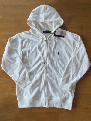 Vit hoodie från Polo Ralph Lauren - Snygg vit hoodie från Polo Ralph Lauren med dragkedja framtill och klassisk huva med dragsko. Tröjan har två fickor fram och den ikoniska mörkblå loggan broderad på bröstet. Tillverkad i mjuk bomull för en skön och avslappnad stil.