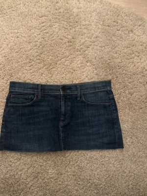 Mörkblå jeanskjol i kort modell - Säljer en mörkblå kort jeanskjol med klassiska fem fickor och bälteshällor. Kjolen har knapp och dragkedja framtill samt snygga sömmar. Perfekt för dig som gillar en enkel men trendig denimlook.