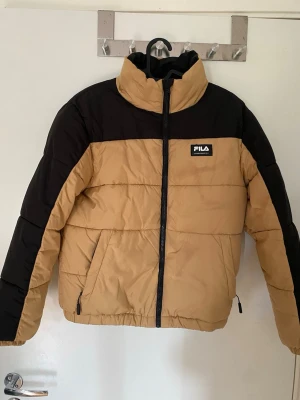 Beige och svart pufferjacka från FILA - Snygg beige och svart pufferjacka från FILA med hög krage och dragkedja framtill. Jackan har två sidofickor med dragkedja och ett broderat FILA-märke på bröstet. Perfekt för kalla dagar och riktigt skön att bära.