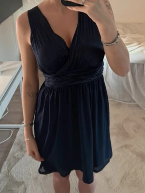 Mörkblå omlottklänning med v-ringning - Snygg mörkblå kortklänning med omlott och djup v-ringning. Klänningen har markerad midja och är ärmlös, vilket ger en elegant och stilren look. Perfekt för dig som vill ha en klassisk och tidlös stil.