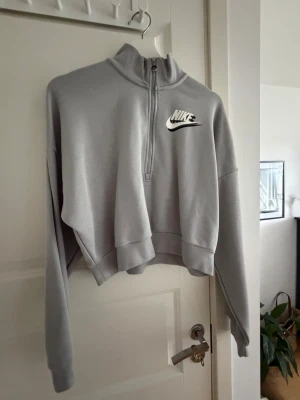 Grå half-zip hoodie från Nike - Snygg grå hoodie från Nike i loose fit-modell med halv dragkedja framtill och hög krage. Tröjan har ett vitt Nike-logotyptryck på bröstet och är tillverkad i mjukt sweatshirtmaterial. Perfekt för en avslappnad och sportig stil.