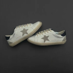 Vita sneakers från Golden Goose - Stilrena vita sneakers från Golden Goose med grå stjärndetalj på sidan och mörkblå häl. Skorna har beige/svart sula och vita skosnören. Tillverkade i läder med klassisk låg profil och diskret branding på sidan. Perfekta för en trendig och avslappnad look.