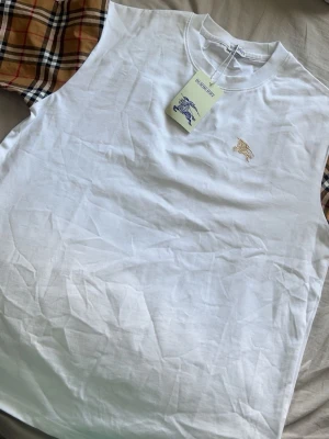 Vit Burberry t-shirt med rutiga ärmar - Säljer en vit t-shirt från Burberry med klassiska beige-rutiga ärmar och guldbroderad logga på bröstet. T-shirten har rund hals och är tillverkad i mjuk bomull. Perfekt för dig som vill ha en stilren men ändå unik look.