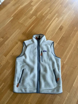 Vit fleeceväst från Patagonia - Mysig vit fleeceväst från Patagonia med hög krage och dragkedja framtill. Västen har två sidofickor och en bröstficka, alla med dragkedja och snygga läderdetaljer. Perfekt lager på lager och riktigt skön att ha på sig under kyliga dagar. Priset kan diskuteras