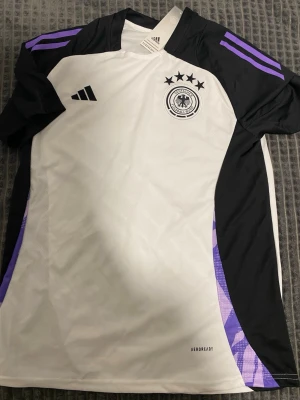 Tysklands matchtröja Adidas vit/svart - Snygg Tyskland matchtröja från Adidas i vitt och svart med lila detaljer på axlarna. Tröjan har fyra stjärnor och landslagets emblem på bröstet samt Aeroready-material som andas. Perfekt för fotbollsträning eller att bära till vardags.
