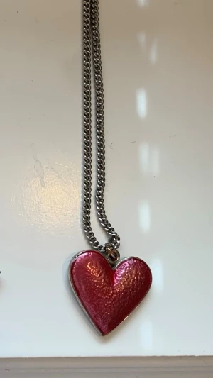 Halsband med rosa hjärthänge - Snyggt halsband med ett glansigt rosa hjärta som hänge. Kedjan är i silverfärgad metall och hjärtat har en skimrande yta som fångar ljuset. Perfekt accessoar för att ge din outfit en romantisk touch.