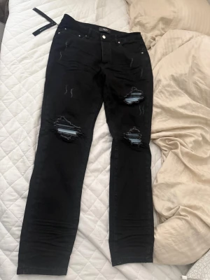 Svarta ripped jeans från Amiri - Säljer ett par svarta jeans från Amiri med slitningar och stora hål på båda knäna. Jeansen har raka ben och klassisk femficksmodell. Materialet är denim och de har coola detaljer med trasiga partier som visar blått tyg under.