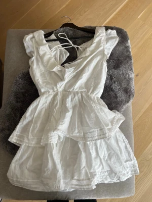 Vit omlottklänning med volang - Söt vit kortklänning i omlottmodell med volangdetaljer och v-ringning. Klänningen har ärmlös design med dekorativa kanter och knytband i ryggen. Perfekt för varma dagar och sommarens alla tillfällen.