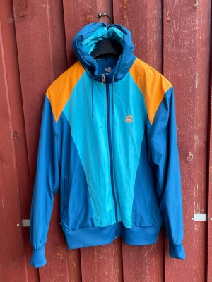 Nike vindjacka - Snygg retro vindjacka med huva från nike. Jackan har en färgstark design i olika blå nyanser och orange detaljer på axlarna. Dragkedja framtill, två sidofickor och resår vid ärmslut och midja. Perfekt för dig som gillar sportig och färgglad stil.
