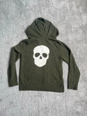 Olivgrön hoodie med döskallemotiv - Cool olivgrön hoodie med stor vit döskalle på ryggen. Hoodien har en klassisk huva och är tillverkad i ett mjukt stickat material som känns skönt mot huden. Perfekt för dig som gillar streetstyle och vill sticka ut lite extra.
