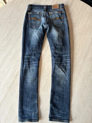 Nudie jeans tvättdesign - Säljer ett par blå slim fit jeans från Nudie Jeans med klassisk femficksmodell och snygga slitningar. Jeansen har kontrastsömmar och broderade bakfickor. Tillverkade i denim med en skön stretch för extra komfort.