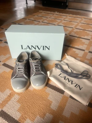 Lanvin DBB1 Patent Cap Toe Sneaker strl 39/40/uk 5 - Sneakers från franska Lanvin i ljusgrå mocka. Ett par stilfulla tillika lediga sneakers tillverkade i mocka. Prydda med den högglansade tåkappan som tillför extra elegans åt skorna och som är signifikant för varumärket. Inköpta på NK Gbg - använda men i fortsatt fint skick, Storlekarna är större så 39 är som en 40. 