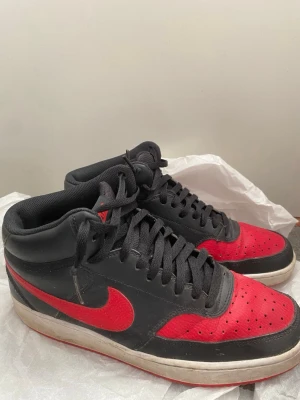 Nike svart-röda sneakers mids - Snygga Nike sneakers i svart och rött med klassisk Swoosh-logga på sidorna. Skorna har svarta skosnören, perforerad tåbox och mellanhögt skaft. Ovandelen är i läder och textil, med vit och röd sula som ger en sportig vibe.