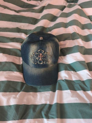 Jeanskeps med broderad detalj - Säljer en snygg jeanskeps med slitna detaljer och broderat mönster framtill. Kepsen har en cool tvättad look och en liten glittrig knapp på toppen. Perfekt för dig som gillar streetstyle och vill sticka ut lite extra.