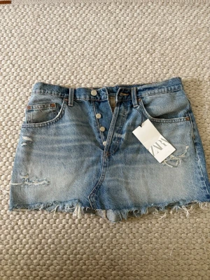 Blå jeanskjol från Zara - Säljer en blå jeanskjol från Zara i klassisk denim med slitna detaljer i strl M. Den är oanvänd därav lappen kvar!