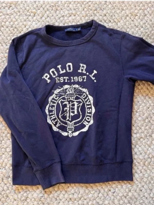 Blå sweatshirt från Polo Ralph Lauren - Mörkblå sweatshirt från Polo Ralph Lauren med stort vitt tryck framtill och rund hals. Tröjan har långa ärmar och ribbade muddar vid ärmslut och nederkant. Perfekt för en avslappnad och sportig look.🙌