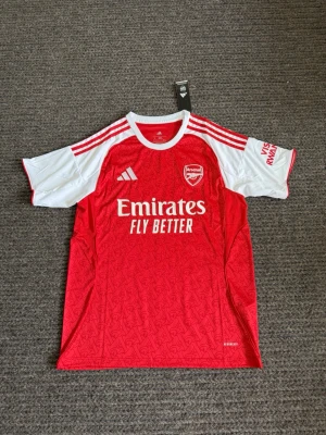 Fotbollströja  - Snygg Arsenal matchtröja från Adidas i rött och vitt med klubbmärke och Emirates Fly Better-tryck på bröstet. Tröjan har vita detaljer på axlar och ärmar samt klassiska Adidas-ränder. Tillverkad i lätt och ventilerande polyester, perfekt för fotboll.
