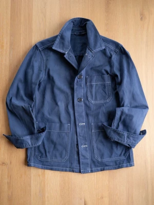 Marinblå overshirt / Chore Jacket med knappar - Stilren marinblå overshirt med tre framfickor och tydliga vita sömmar. Jackan har klassisk krage, knäppning framtill och är tillverkad i bomull. Perfekt lager-på-lager-plagg för en avslappnad look. Lite color bleed som kan ses på bilderna och fin patina. 