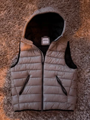 Beige dunväst med huva från Bershka - Snygg beige dunväst från Bershka med svart innerfoder och dragkedja framtill. Västen har två snedställda fickor med dragkedja och en stor huva. Perfekt för lager på lager och streetwear-vibe.