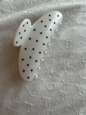 Vit hårklämma med svarta prickar - Säljer en stor vit hårklämma i plast med svarta prickar. Klämman har en klassisk form med rejäla taggar och metallfjäder för bra grepp. Perfekt accessoar för att snabbt fixa frisyren och ge en trendig touch till din look. Secondhand 