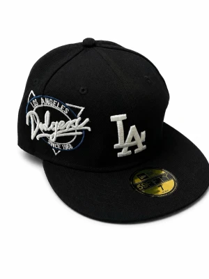 Svart LA Dodgers keps New Era - Säljer en svart keps från New Era med broderad LA-logga i vitt framtill och Los Angeles Dodgers-märke på sidan. Klassisk 59FIFTY-modell med platt skärm och guldfärgad etikett på skärmen. Perfekt för dig som gillar streetwear och sportig stil.
