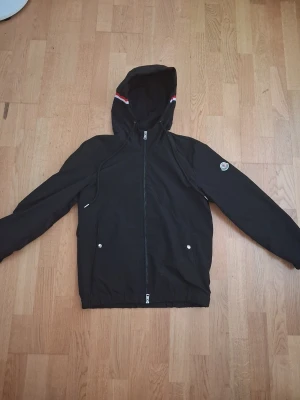 Svart vindjacka från Moncler - Svart vindjacka från Moncler med huva och dragkedja framtill. Jackan har två fickor med tryckknappar och en Moncler-logga på ena ärmen. Detaljer med röd och vit rand på huvan. Perfekt för blåsiga dagar.
