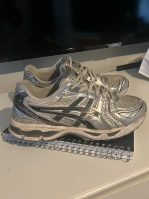 Silvriga Asics sneakers - Säljer ett par riktigt snygga silvriga Asics sneakers med mesh och syntetmaterial. Skorna har klassisk låg profil, vita och grå detaljer samt den ikoniska Asics-loggan på sidan. Perfekta för dig som gillar sportig och trendig stil.