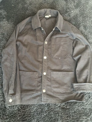 Svart overshirt från Fristads - Passar strl 158 Stilren svart overshirt från Fristads med tre stora utanpåfickor och klassisk krage. Overshirten har tryckknappar framtill och är gjord i ett slitstarkt bomullstyg. Perfekt lager-på-lager-plagg för en avslappnad look.