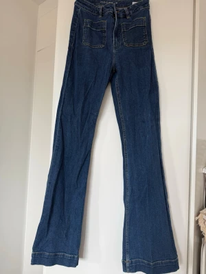 Blå utsvängda jeans från Bikbok - Säljer ett par blå jeans från Bikbok med utsvängda ben och hög midja. Jeansen har två stora fickor framtill och klassisk femficksdesign. Perfekta för dig som gillar retrostil och vill ha ett par bekväma och snygga jeans i garderoben.