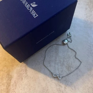 Swarovski silverarmband med kristaller - Snyggt och elegant armband från Swarovski i silverfärgad metall med en tunn kedja och ett gnistrande dekorelement i form av en rosett med små klara kristaller. Perfekt för dig som gillar stilrena smycken med lite extra bling.