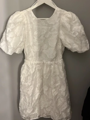 Vit kortklänning med puffärm och snörning - Supersöt vit kortklänning med puffärmar och strukturmönster i tyg. Klänningen har öppen rygg med snörning och markerad midja. Perfekt för dig som vill ha en romantisk och trendig look. Materialet känns lätt och har en blommig struktur.
