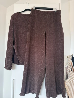 Brunt sett från Nakd  - Säljer ett par bruna plisserade leggings med vida ben och elastisk midja. Materialet är mjukt och stretchigt med en snygg struktur. Perfekta för dig som gillar en avslappnad men trendig stil. Matcha med en oversized tröja i samma stil för en cool look.