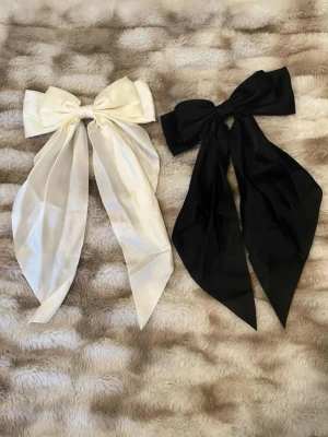 Stora Hårrosetter i Svart och Vitt - Två stora hårrosetter i satinliknande material, en i svart och en i vitt. Båda har långa band och klassisk rosettform som ger en elegant och trendig look. Perfekta för att piffa upp frisyren och skapa en statement-accessoar. Använt endast den svarta en gång, så de är stort sett helt nya.