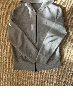 Grå hoodie från Polo Ralph Lauren - Klassisk grå hoodie med dragkedja från Polo Ralph Lauren. Tröjan har huva, två fickor framtill och den ikoniska lilla loggan broderad på bröstet. Tillverkad i mjukt sweatshirtmaterial som är perfekt för en avslappnad stil.