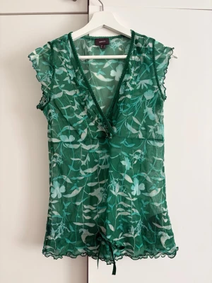 Grön blommig omlottopp från Mexx - Snygg grön omlottopp från Mexx med blommigt mönster och transparent meshmaterial. Toppen har v-ringning, volangkantade ärmar och knytband nedtill. Perfekt för dig som gillar en lätt och luftig stil. Made in Spain.
