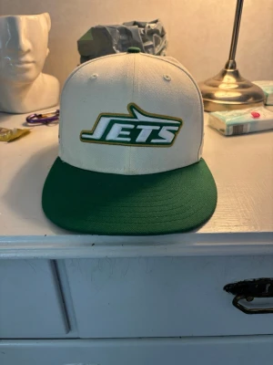 Jets Fitted  - Säljer en snygg Jets keps med vit krona och grön skärm. Framtill finns ett broderat Jets-logotyp i grönt och vitt med guldkant. På sidan står det 'Super Bowl III' broderat i grönt och guld. Klassisk snapback-modell, perfekt för dig som gillar sportig stil.