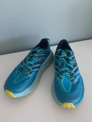 Blå Hoka sneakers med gula detaljer - Snygga blå sneakers från Hoka med gula detaljer på tå och sula. Skorna har en chunky sula och mönstrad yttersula för extra grepp. Ovandelen är i syntetmaterial med randiga inslag och matchande blå-gula skosnören. Perfekta för dig som gillar sportig stil.