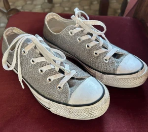 Silvriga Converse låga sneakers - Säljer ett par silvriga Converse sneakers med klassisk vit tåhätta och vita snören. Skorna har en glittrig textilöverdel och vit sula med svart rand. Perfekta för dig som vill sticka ut lite extra i vardagen.