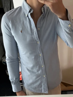 Ljusblå skjorta från Ralph Lauren - Klassisk ljusblå skjorta från Ralph Lauren med button-down krage och broderad logga på bröstet. Skjortan har lång ärm, vita knappar och är tillverkad i mjuk bomull. Perfekt för en clean och stilren look.