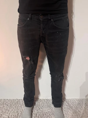 Dondup George Jeans - Säljer ett par svarta skinny jeans med slitningar och hål på båda benen. Jeansen har fem fickor, låg midja och en tvättad look med små färgstänk. Perfekta för dig som gillar en edgy och trendig stil.