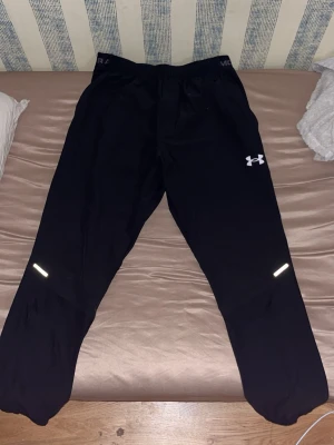 Svarta träningsbyxor Under Armour - Säljer ett par svarta träningsbyxor från Under Armour med resår i midjan och reflexdetaljer på benen. Byxorna har en sportig passform och är tillverkade i ett lätt och snabbtorkande material, perfekta för löpning eller gym.
